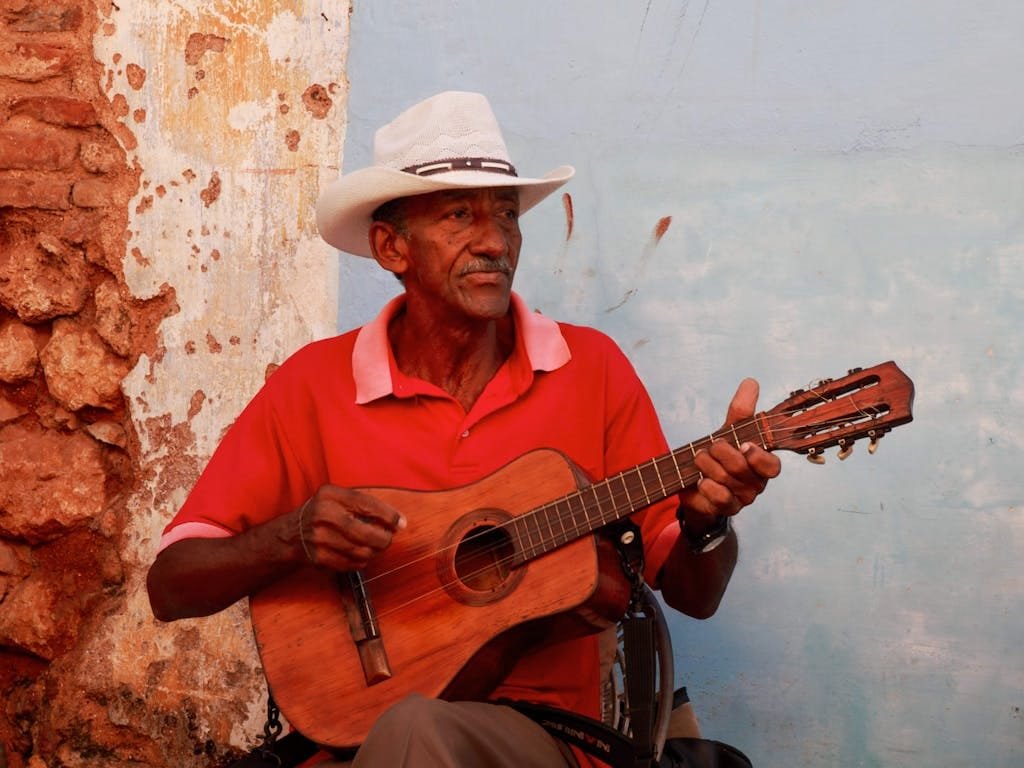 A senior man in a cowboy hat plays a guitar against a rustic wall, creating a candid musical moment.